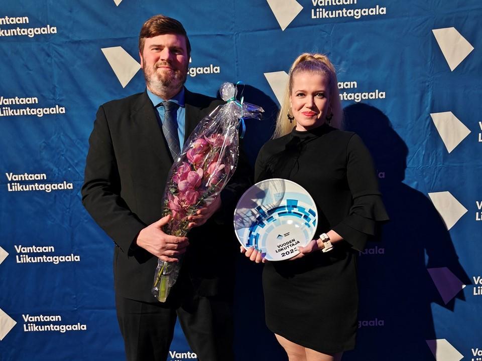 Kaksi henkilöä Vantaan Liikuntagaalan lakanan edessä kukkapuska ja palkinto käsissä.